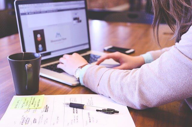 Mujer escribiendo en computadora sobre mesa en la que hay teléfono, taza y papel con anotaciones